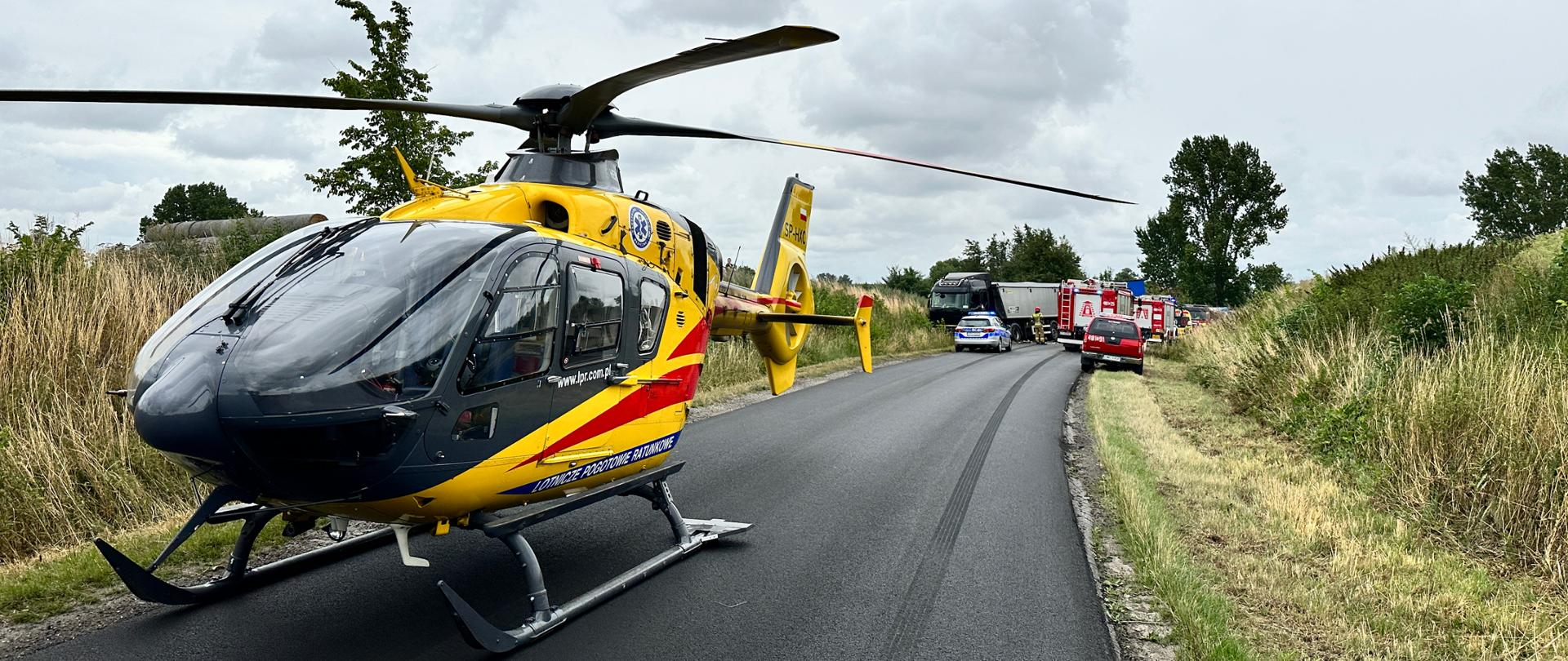 Po lewej stronie znajduje się śmigłowiec Lotniczego Pogotowia Ratunkowego. W oddali widać stojący na poboczu samochód ciężarowy z naczepą oraz samochód policyjny. Po prawej stronie w oddali stoją samochody straży pożarnej. 