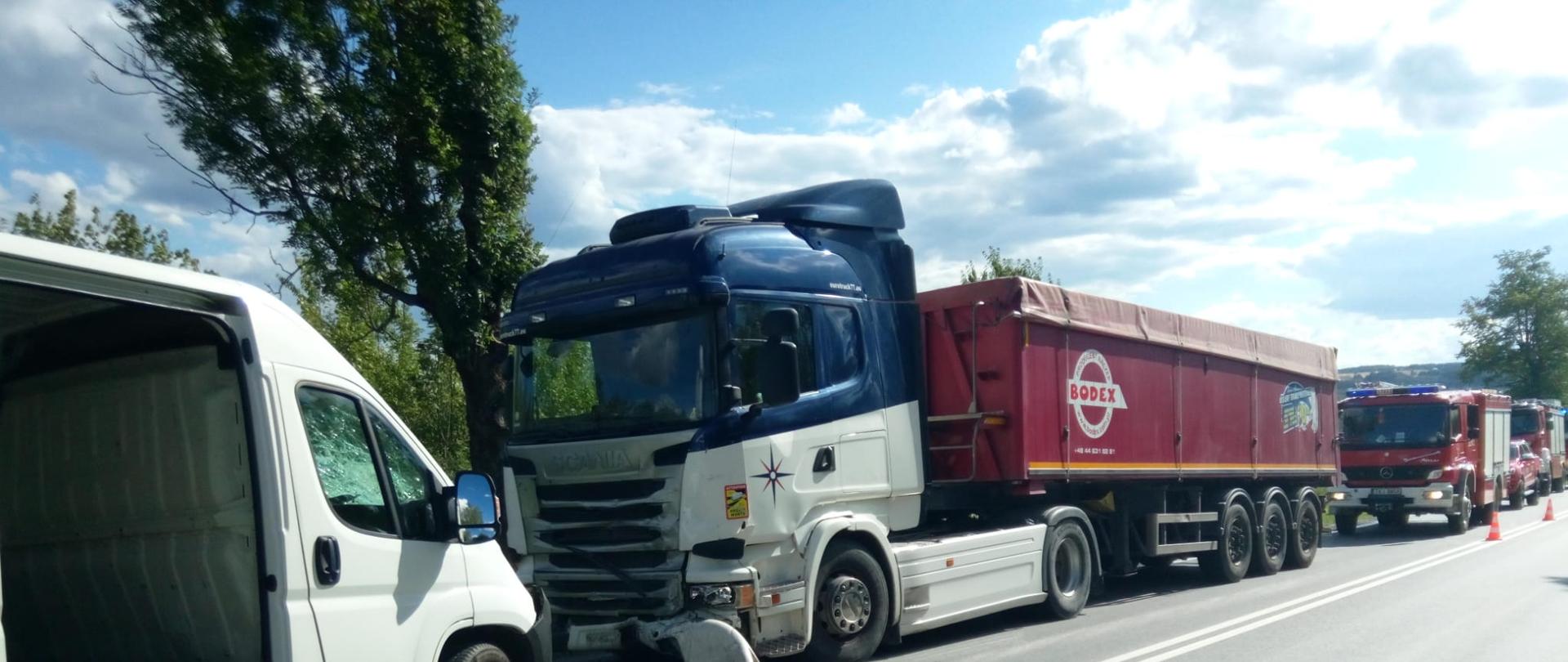 Zdjęcie przedstawia miejsce wypadku: z lewej strony dwa uszkodzone auta: bus i ciągnik siodłowy, a za nimi samochody pożarnicze.
