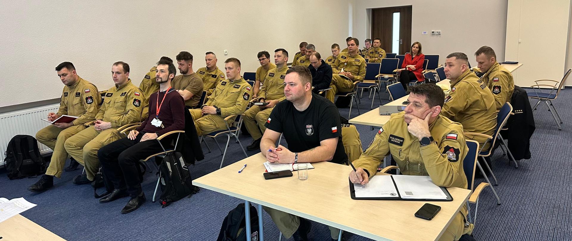 Ujęcie sali konferencyjnej z innej perspektywy – strażacy PSP w mundurach koloru khaki oraz jedna kobieta w czerwonej marynarce słuchają prelekcji. Niektórzy trzymają notesy i długopisy, a na ścianie wisi zegar.