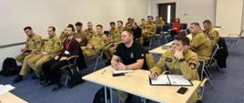 Ujęcie sali konferencyjnej z innej perspektywy – strażacy PSP w mundurach koloru khaki oraz jedna kobieta w czerwonej marynarce słuchają prelekcji. Niektórzy trzymają notesy i długopisy, a na ścianie wisi zegar.