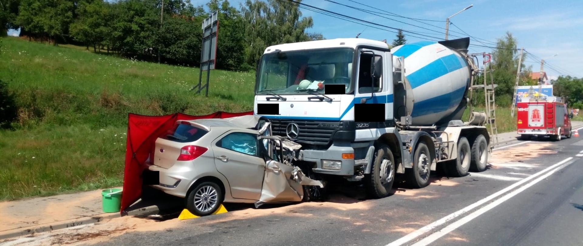 Na zdjęciu widać samochód ciężarowy typu betoniarka do przewozu betonu oraz wrak samochodu osobowego po zderzeniu czołowym. W tle samochód ratowniczo gaśniczy OSP oraz drzewa.