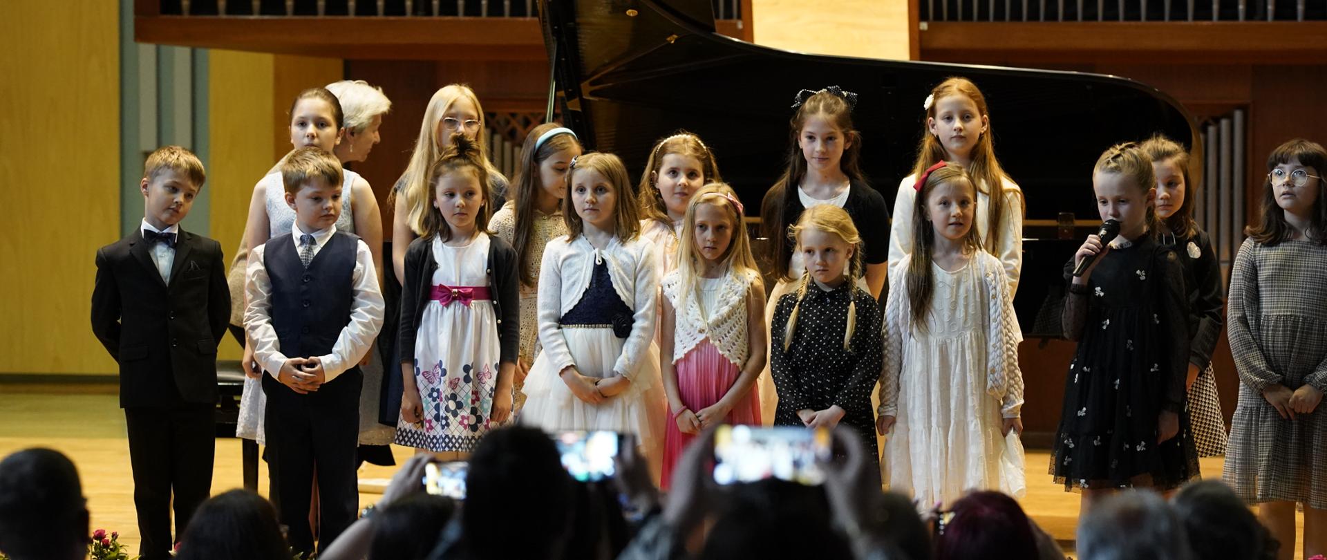 Młodzi pianiści stoją w dwóch rzędach na scenie w sali koncertowej, w tle organy oraz fortepian koncertowy. Na pierwszym planie rodzice robiący zdjęcia telefonami komórkowymi