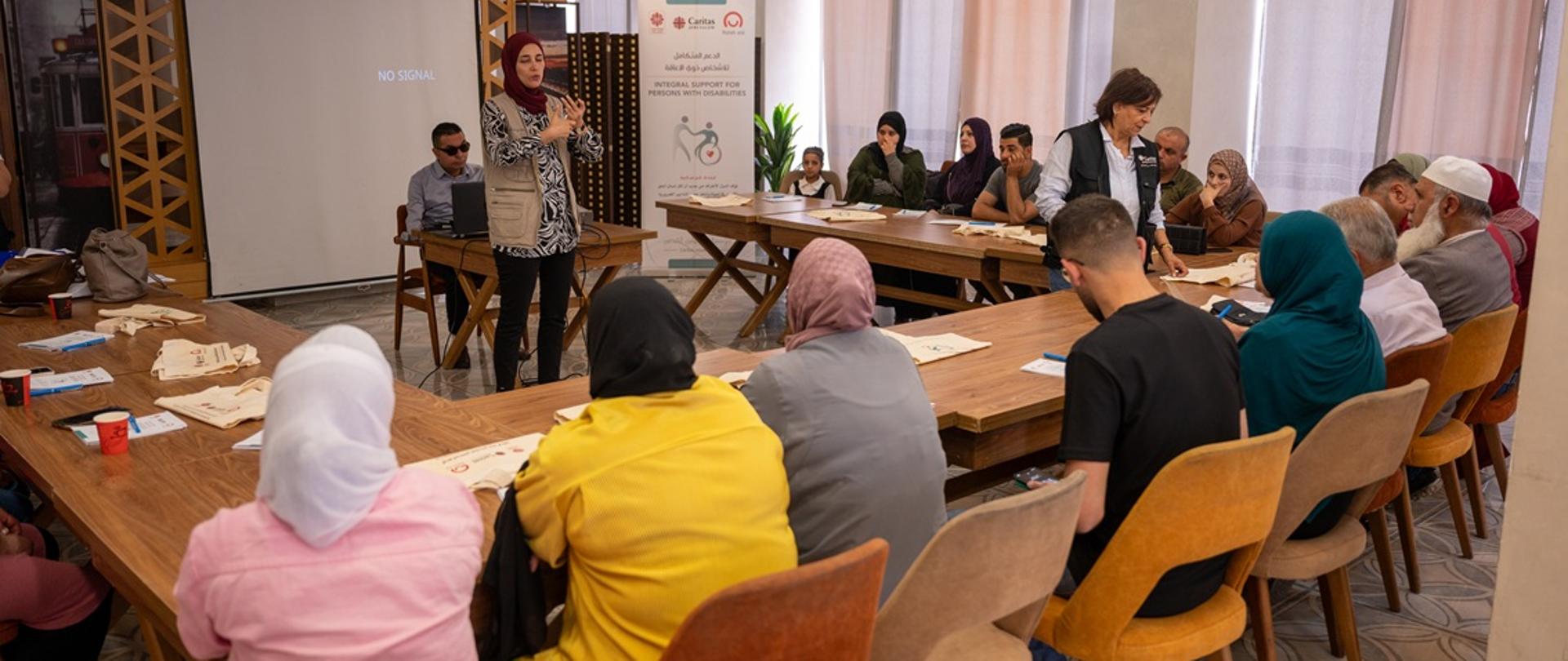 Grupa ludzi, kobiet i mężczyzn, siedzi wokół ustawionego w podkowę stołu, stojąca kobieta w chuście prowadzi spotkanie