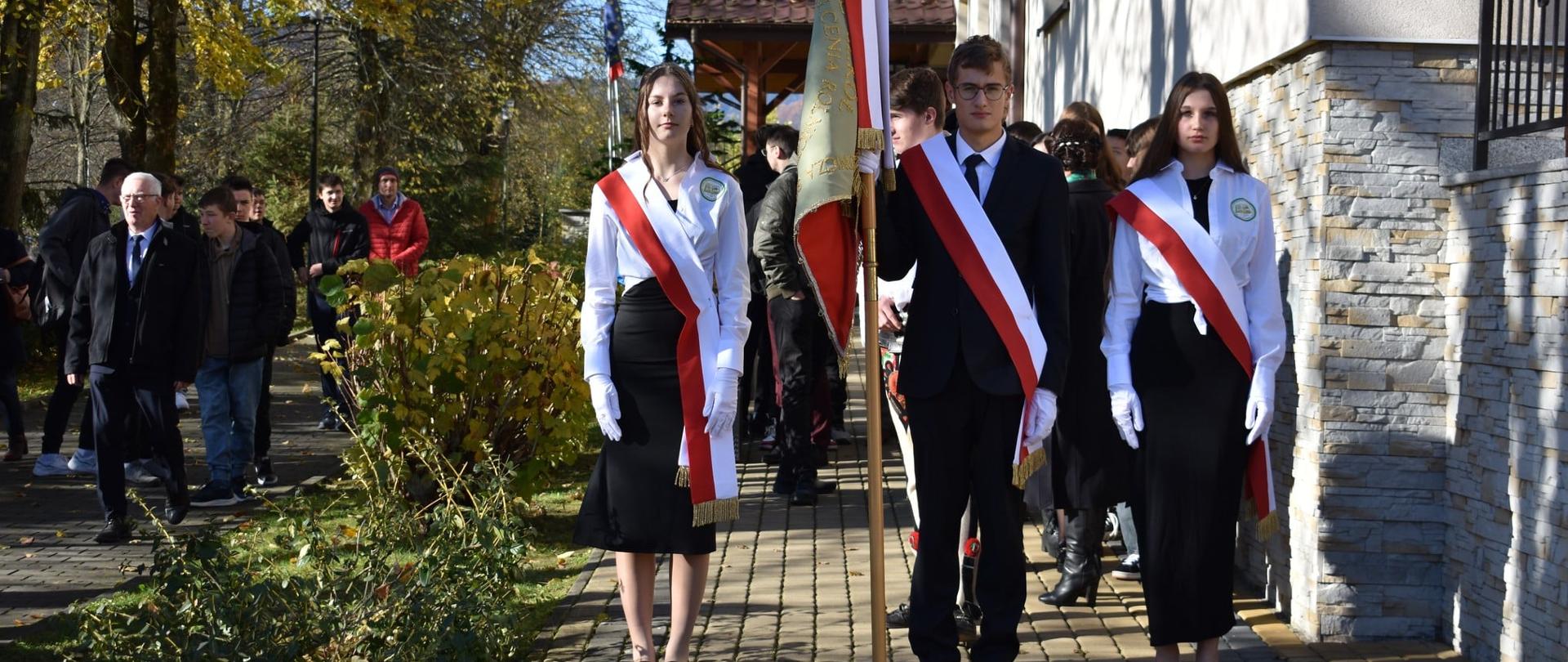 Poczet sztandarowy szkoły (dwie dziewczyny i chłopak) w galowych strojach z biało-czerwonymi szarfami stoi przed budynkiem. W tle widać gości i jesienne drzewa.