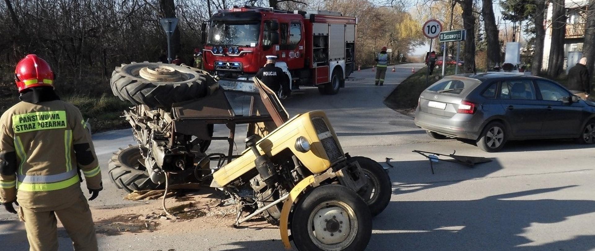 Uszkodzony ciągnik rolniczy na drodze. Tyłem odwrócony strażak. W tle samochód gaśniczy.