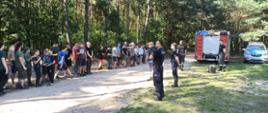 na pierwszym planie dwóch policjantów dalej dwóch strażaków dalej stoi samochód strażacki oraz policyjny na drodze polnej trwa zbiórka uczestników obozu - próbna ewakuacja za nimi drzewa