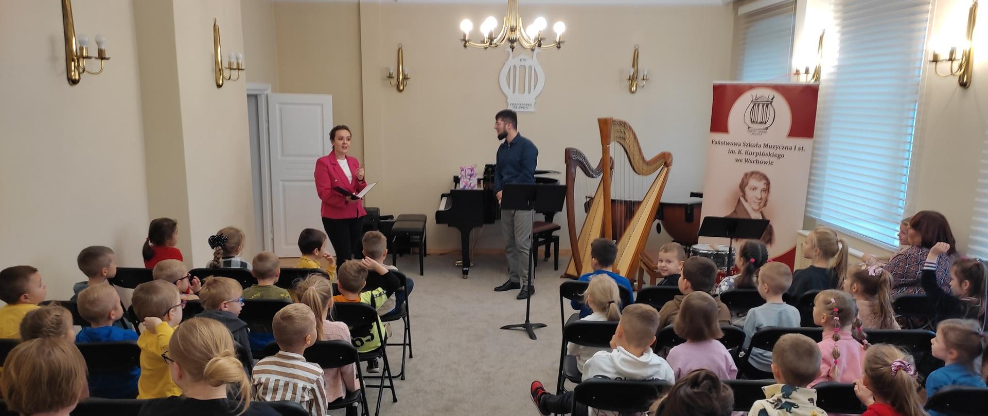 Na zdjęciu widoczni są nauczyciele szkoły prowadzący koncert: pani Dominika Stachowicz oraz p. Krystian Klej. Tyłem do obiektywu siedzą dzieci oraz nauczyciele z przedszkoli. W tle widoczny fortepian, dwie harfy oraz baner z wizerunkiem patrona i nazwą szkoły.