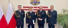 Powierzenie pełnienia obowiązków na stanowisku komendanta miejskiego PSP w Szczecinie st. bryg. Robertowi Tarczyńskiemu. Na zdjęciu widoczni 4 mężczyźni w mundurach wyjściowych, w tle flagi Polski, godło Polski oraz napis "Komenda Wojewódzka PSP w Szczecinie" 