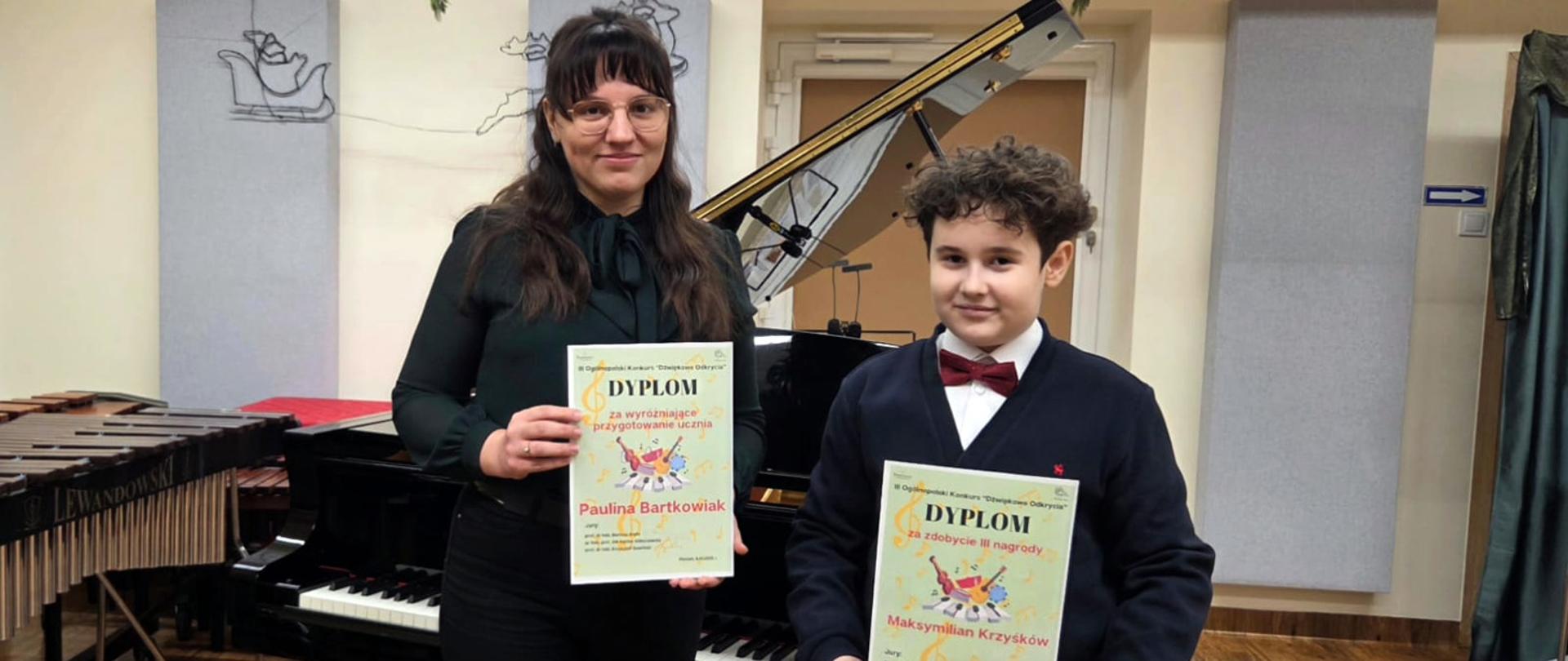 Nauczycielka i uczeń stoją przed fortepianem, oboje w rękach trzymają dyplomy.