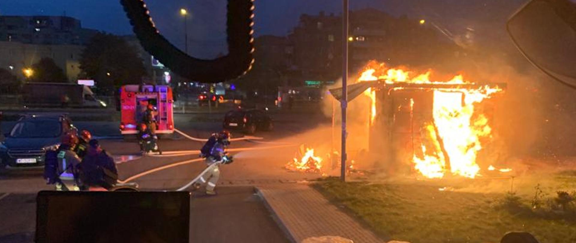 W centralnej części zdjęcia widać strażaka podającego prąd wody w natarciu na rozwinięty pożar kiosku. Z lewej strony zdjęcia widać czerwony samochód ratowniczo-gaśniczy.