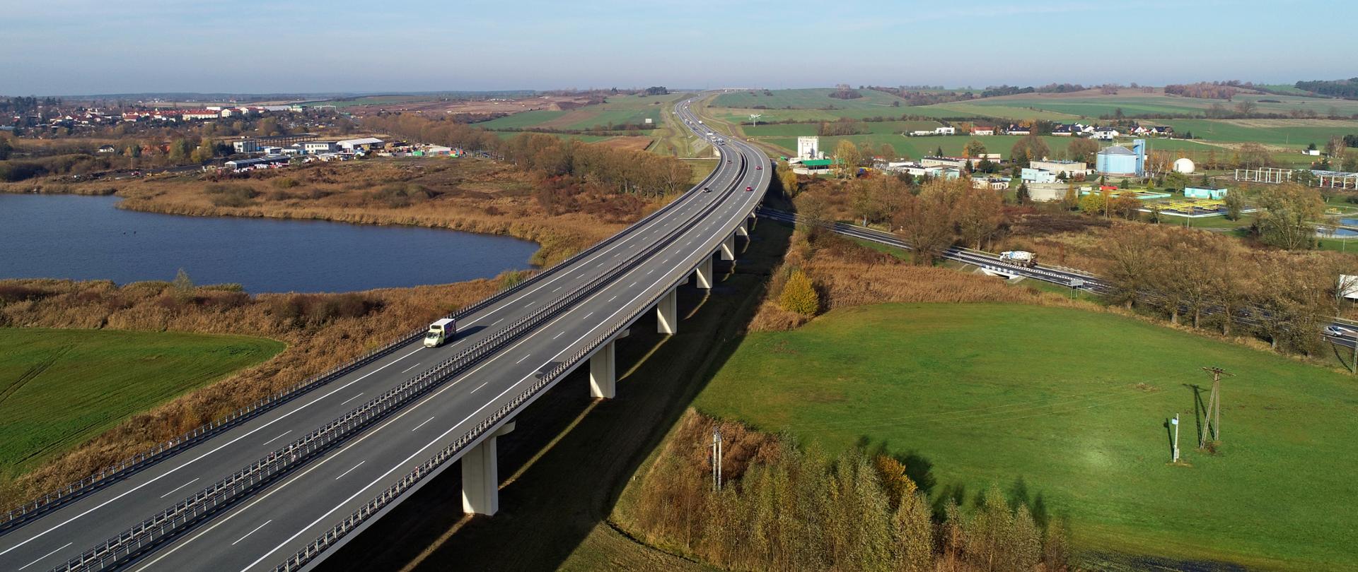 Droga dwujezdniowa biegnąca po estakadzie. Po lewej stronie małe jezioro, po prawej łąka. W oddali zabudowania miejskie.