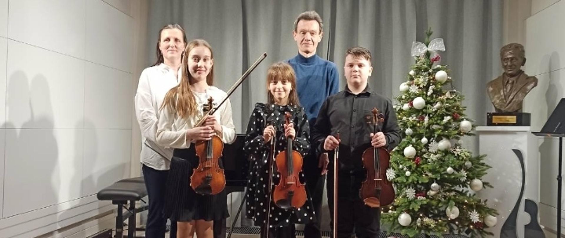 Uczniowie ze skrzypcami stoją na scenie. Po prawej stronie stoi nauczyciel, a po lewej akompaniatorka. Z prawej strony stoi ubrana choinka, a obok popiersie z patronem szkoły. W tle widać zasunięte kotary w szarym kolorze.