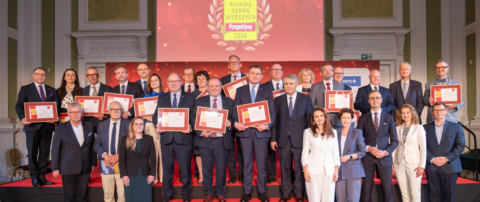 Zdjęcie zbiorowe, na podwyższeniu z przodu sali na czerwonym dywanie stoi grupa osób trzymających dyplomy w czerwonych ramkach, nad nimi na wielkim ekranie napis Ranking Szkół Wyższych Perspektywy 2026.