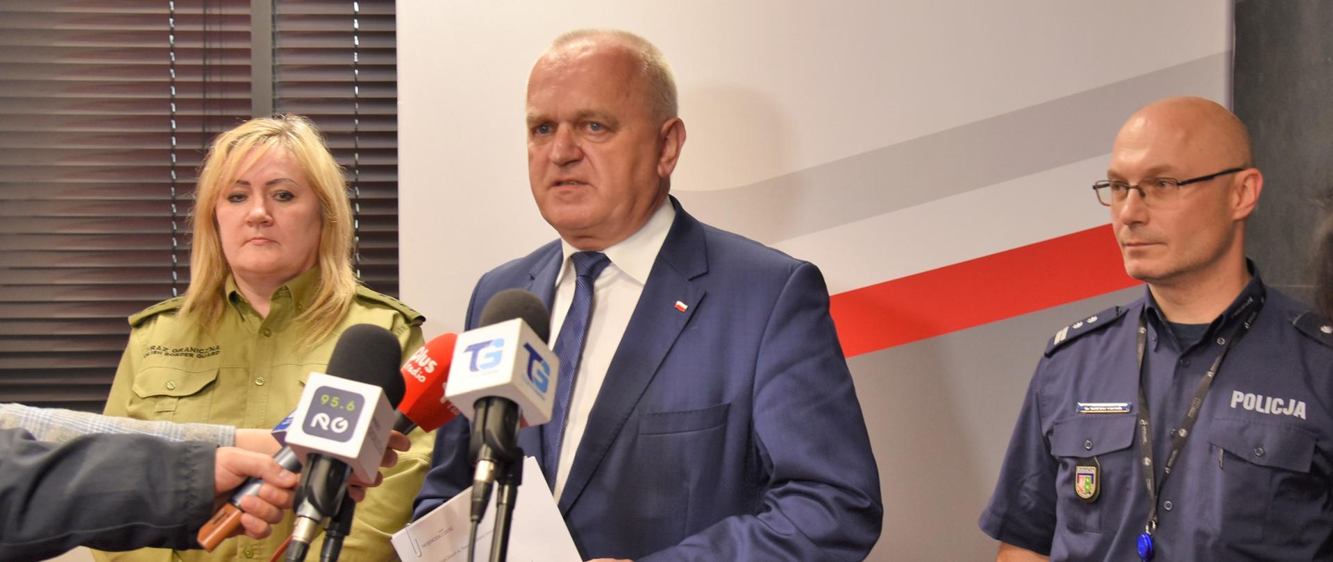 NA tle ścianki promocyjnej urzędu, przed mikrofonami na statywie stoi wojewoda, naczelnik prewencji policji i koordynator Straży Granicznej. Po ich prawej stronie stoi dyrektor wydziału polityki społecznej LUW 