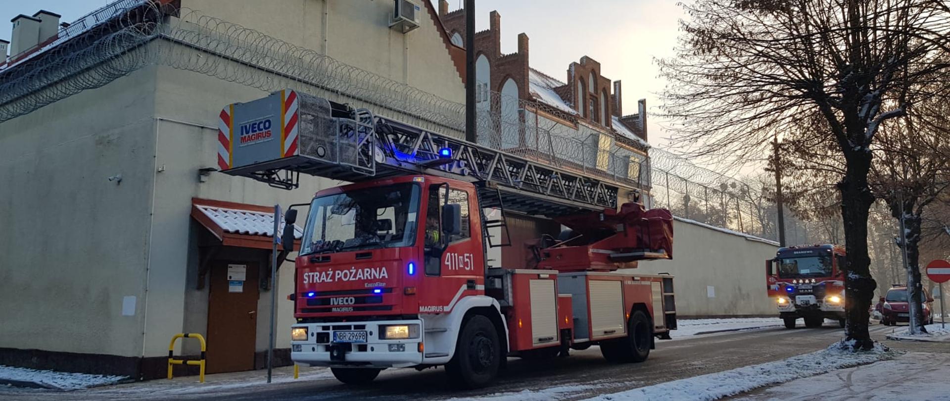 Przy dużym budynku stoją trzy samochody strażackie. Pierwszy samochód drabina strażacka. Pogodnie, biało od śniegu.