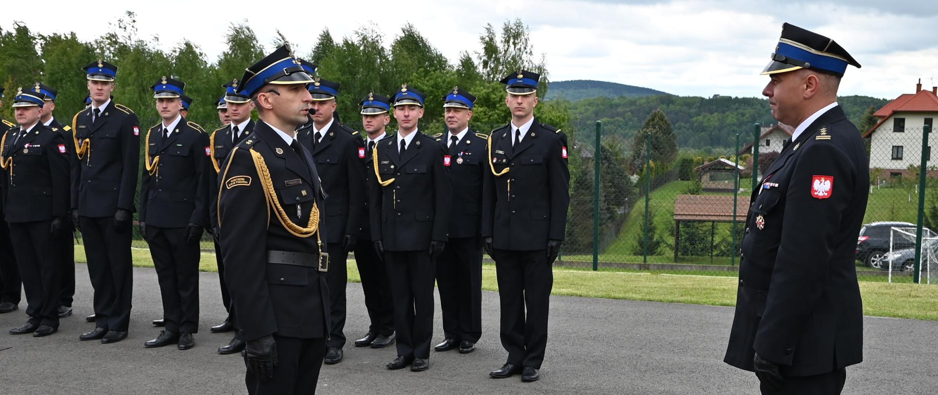 Dwóch strażaków w galowych mundurach salutuje sobie nawzajem podczas obchodów Dnia Strażaka. Za nimi w szyku stoją inni strażacy, wszyscy w czarnych mundurach galowych z żółtymi sznurami. W tle widoczne są budynki mieszkalne, zaparkowane samochody oraz zieleń, co wskazuje na plenerową ceremonię.