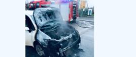 Zdjęcie zrobione na tle samochodu strażackiego. Na zdjęciu spalone auto osobowe, w którym spaleniu uległa komora silnika