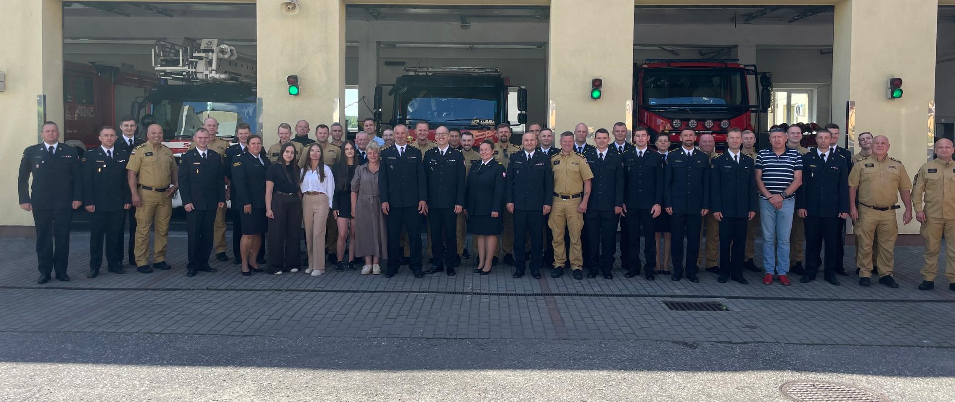 Duża grupa strażaków w ciemnych mundurach wyjściowych, piaskowych mundurach służbowych oraz cywilów pozuje do wspólnego zdjęcia przed budynkiem komendy straży pożarnej, w którego boksach garażowych stoją wozy bojowe.