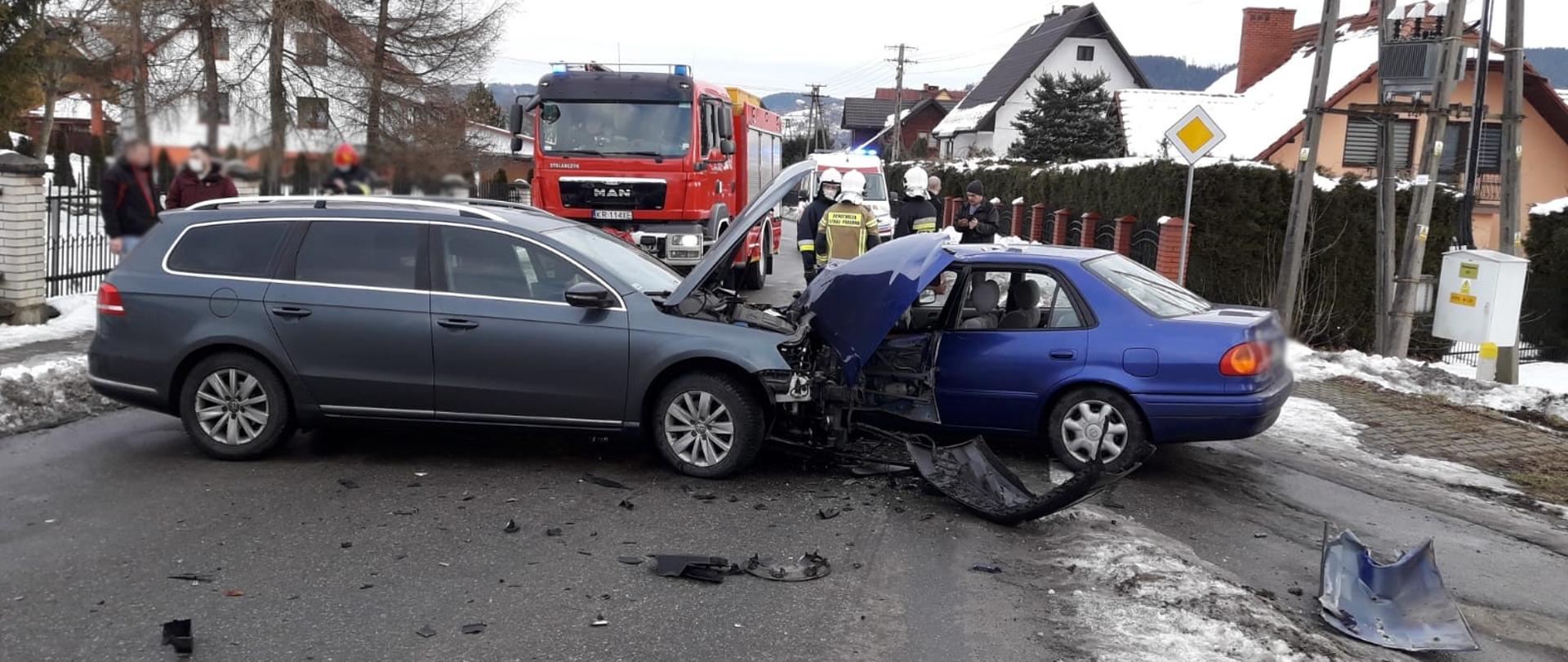 Dwa samochody osobowe na drodze po zderzeniu. W tle samochód ratowniczo-gaśniczy oraz strażacy prowadzący działania.