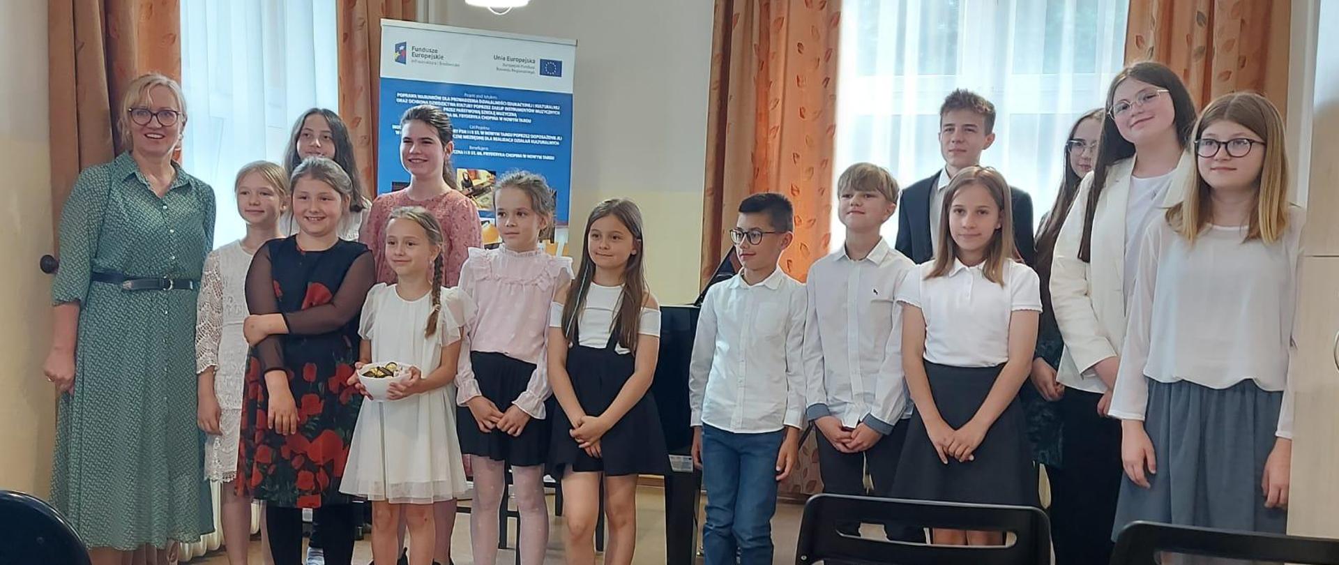 Dzieci stoją w sali kameralnej w uroczystych strojach. Po lewej stronie nauczyciel. Za dziećmi duże okna z długimi zasłonami. Przed dziećmi czarne krzesła. 