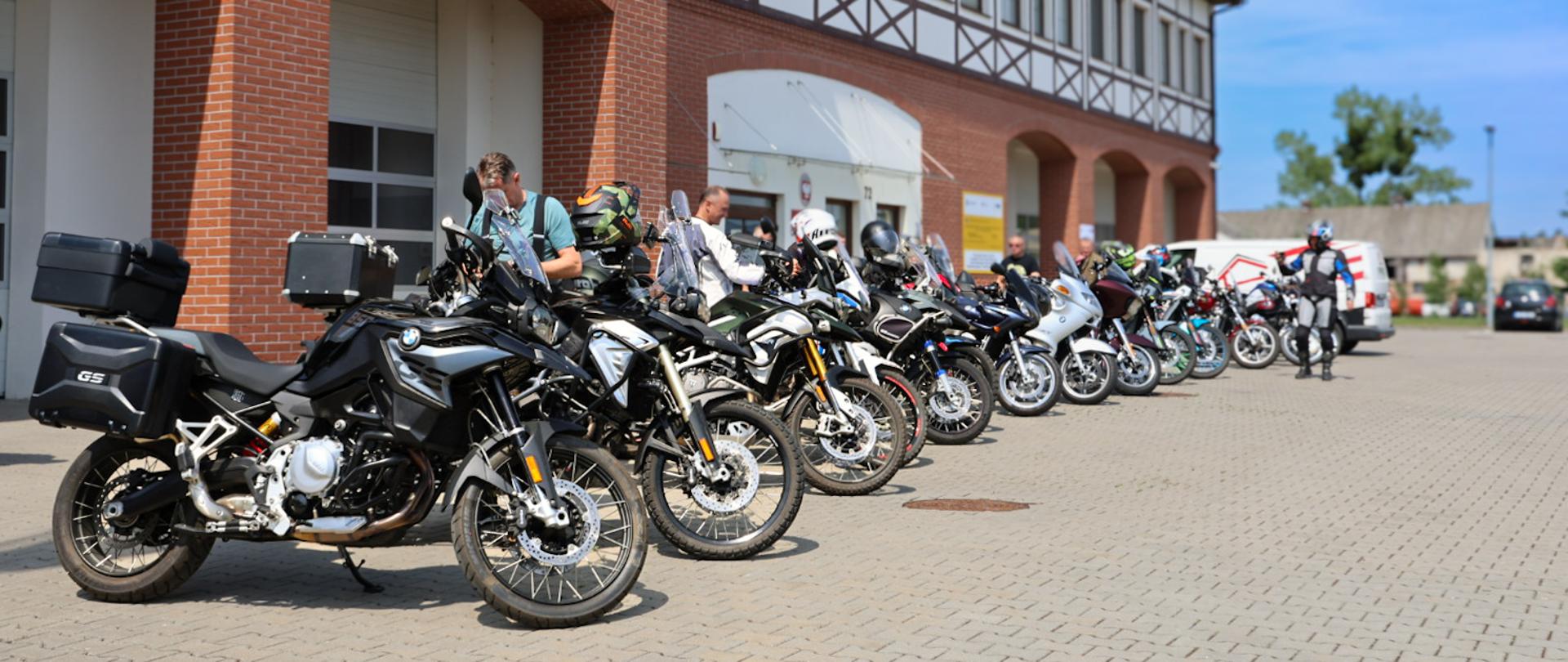 Długi rząd motocykli turystycznych (głównie marek BMW z serii GS) stoi zaparkowany przed budynkiem Ośrodka Szkolenia Państwowej Straży Pożarnej w Łubiance. Przy maszynach widać motocyklistów szykujących się do drogi. Budynek charakteryzuje się architekturą z czerwonej cegły i elementami muru pruskiego. W tle panuje słoneczna pogoda z bezchmurnym, niebieskim niebem.