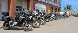 Długi rząd motocykli turystycznych (głównie marek BMW z serii GS) stoi zaparkowany przed budynkiem Ośrodka Szkolenia Państwowej Straży Pożarnej w Łubiance. Przy maszynach widać motocyklistów szykujących się do drogi. Budynek charakteryzuje się architekturą z czerwonej cegły i elementami muru pruskiego. W tle panuje słoneczna pogoda z bezchmurnym, niebieskim niebem.