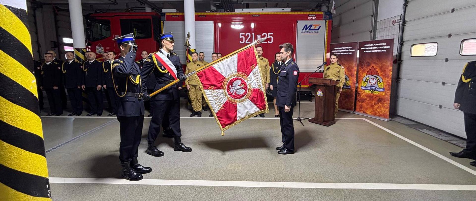 w garażu jednostki starszy brygadier Krzysztof Kowalski stoi na przeciwko pocztu sztandarowego , za nimi w dwuszeregu stoją strażacy i dalej samochód gaśniczy