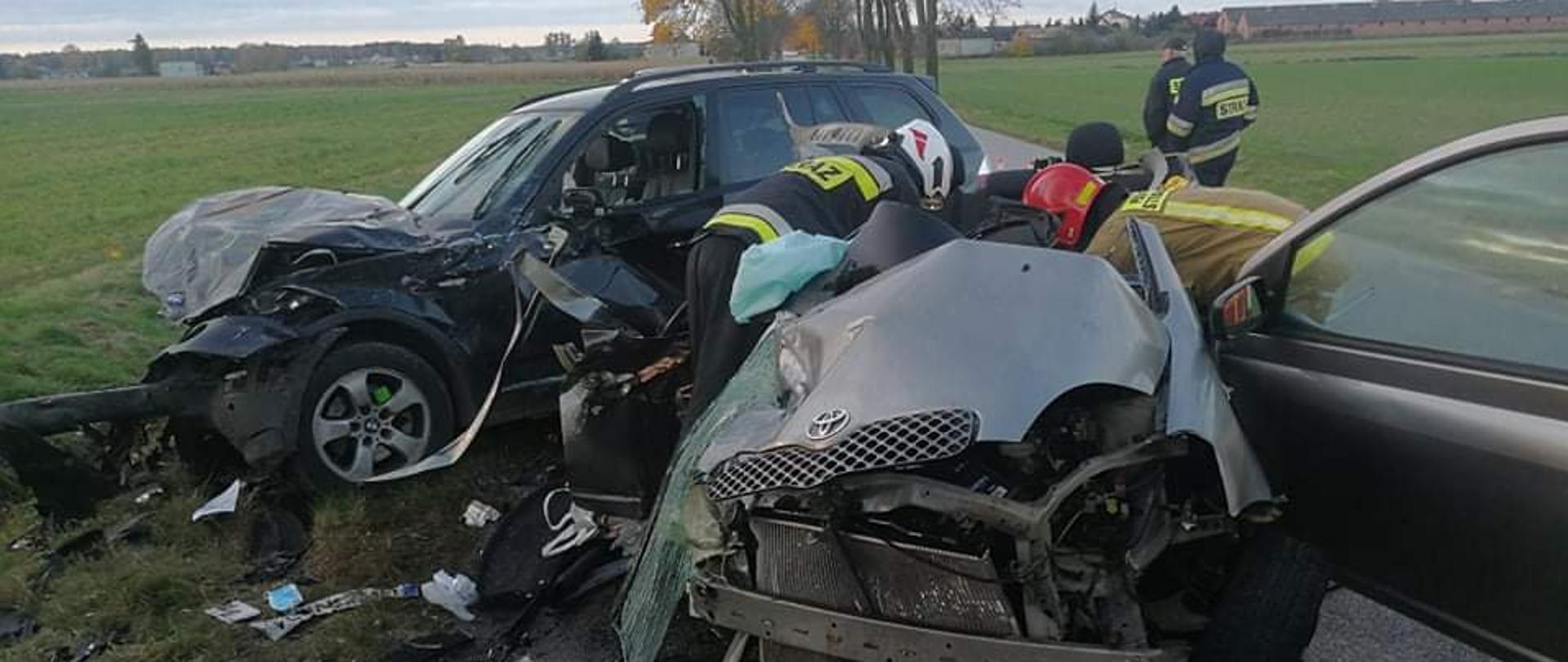 Zdjęcie przedstawia dwa samochody osobowe zdeformowane podczas wypadku. W pobliżu pojazdu rozrzucone elementy karoserii. We wraku pojazdu pracują ratownicy PSP i OSP. 