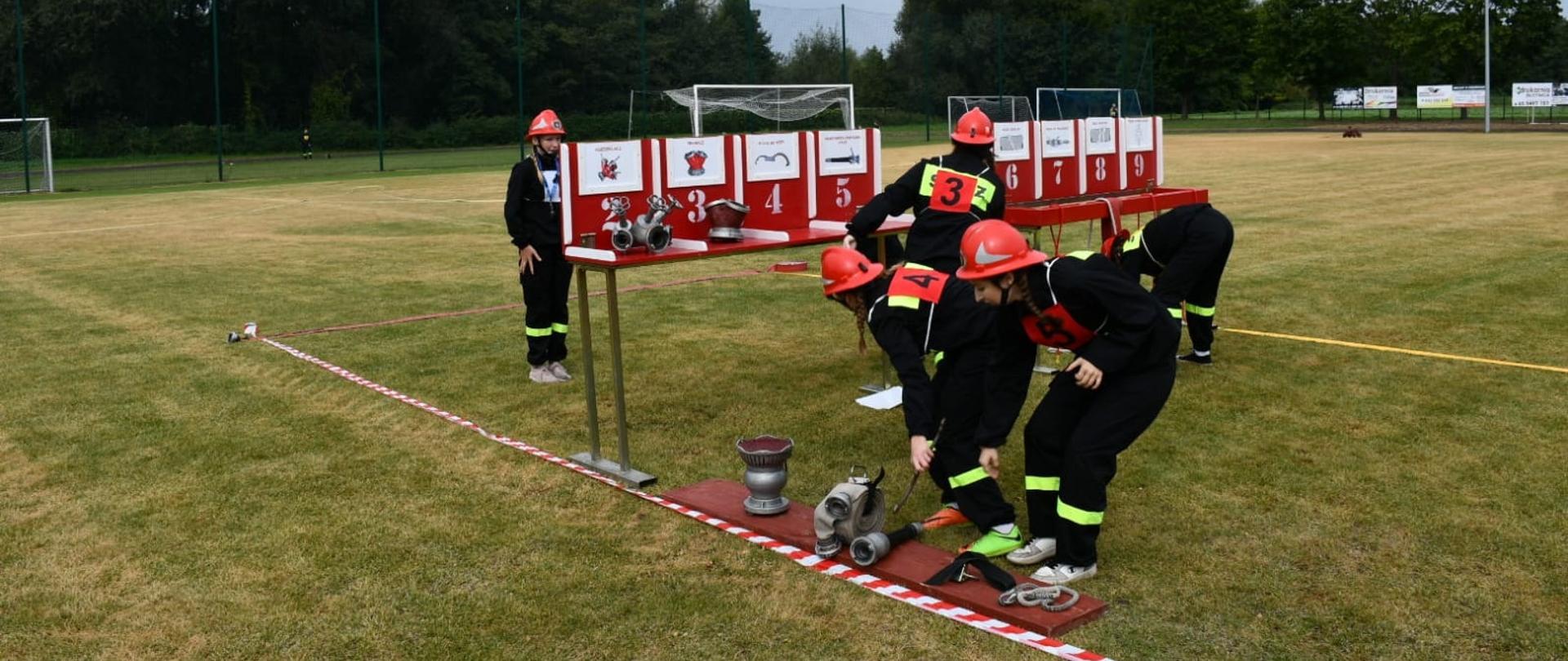 Zdjęcie przedstawia członków Młodzieżowej Drużyny Pożarniczej w czarnych dresach z żółtymi taśmami odblaskowymi, czerwonych hełmach oraz numerach startowych na plecach i piersiach, wykonujących konkurencję rozwinięcia bojowego według regulaminu CTIF na trawiastym boisku sportowym. W centralnej części kadru znajduje się stanowisko przyrządów – czerwono-biały stół z przegrodami oznaczonymi numerami od 1 do 9 i rysunkami sprzętu, przy którym młodzież realizuje zadanie polegające na prawidłowym przyporządkowaniu i obsłudze wyposażenia. Na podeście oraz bezpośrednio na trawie wyznaczonej biało-czerwoną taśmą leży armatura wodna, w tym rozdzielacz, prądownice oraz pożarnicze węże, które zawodnicy podłączają w trakcie próby. Całość odbywa się na boisku otoczonym zielenią, z widoczną w tle bramką sportową, oświetleniem oraz linią drzew pod zachmurzonym niebem.