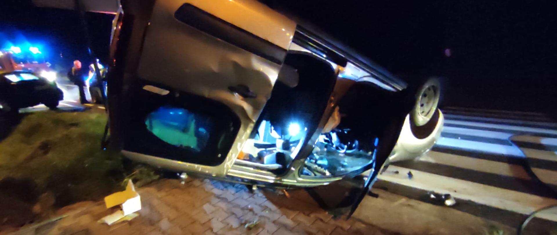 Zdjęcie obrazuje samochód osobowy marki Citroen leżący na dachu z powodu uderzenia jakie otrzymał od samochodu BMW. Drzwi pojazdu są uszkodzone i powgniatana jest karoseria. Obok pojazdu znajduje się przejście dla pieszych. Dookoła widać porozrzucane elementy samochodów . W tle widać światła błyskowe samochodu pożarniczego oraz rozbity samochód BMW i stojącego przy nim policjanta przesłuchującego jednego z uczestników zdarzenia.