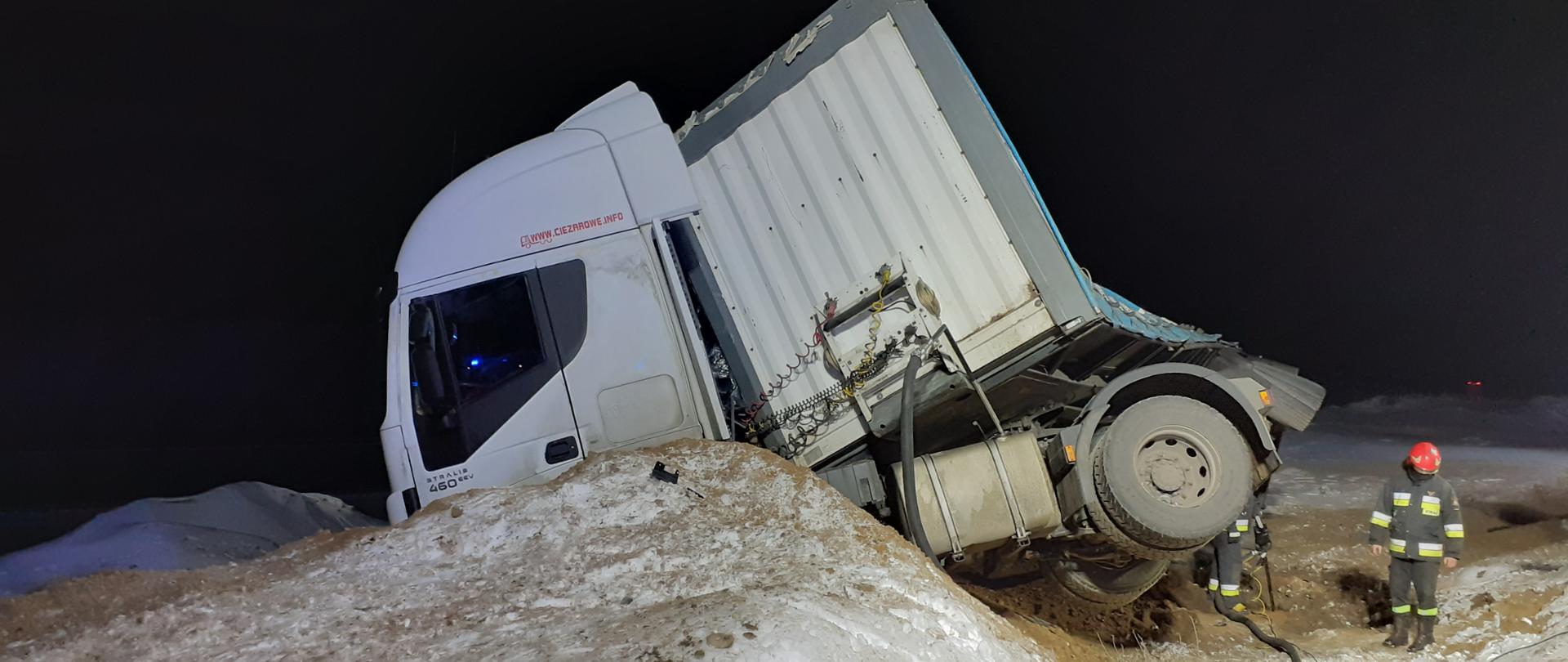 Na zdjęciu widać samochód ciężarowy z pochyloną naczepą. Przed samochodem osobowym widoczna jest hałda piasku pokryta śniegiem. W prawej części zdjęcia widać strażaków prowadzących działania ratownicze.