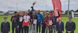 Grupowe zdjęcie zwycięzców na podium, po lewo i prawo stoją funkcjonariusze PSP w mundurach