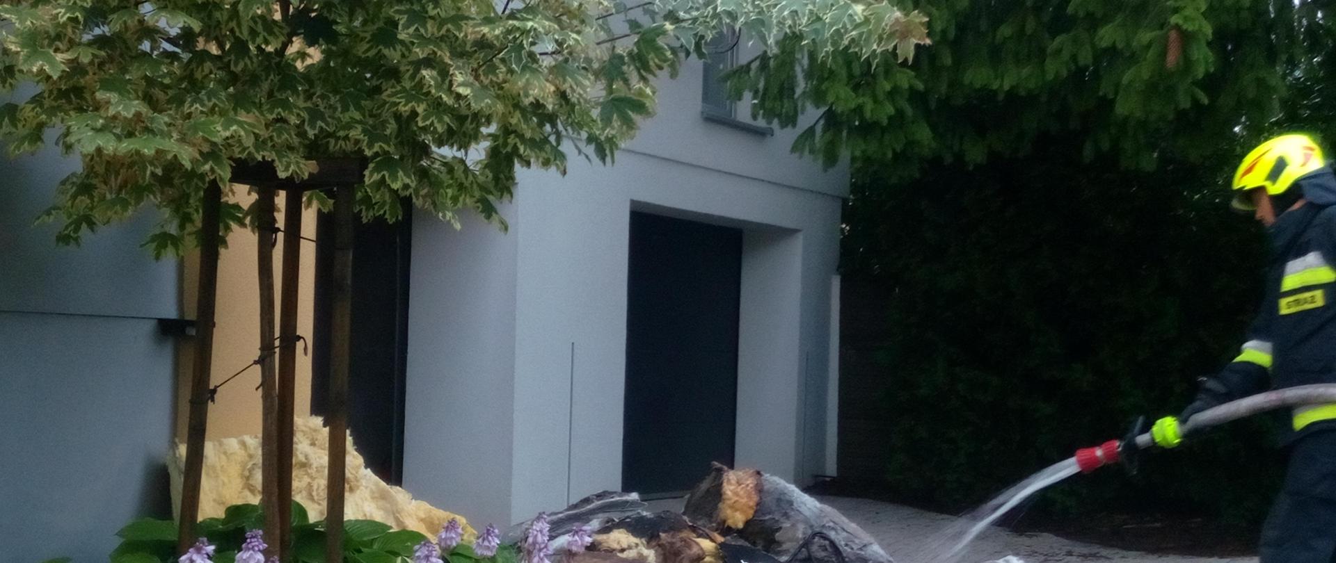 Zdjęcie przedstawia budynek w Kostomłotach, gdzie wybuchł pożar od uderzenia pioruna. Przy ścianie leży pryzma nadpalonej wełny mineralnej przelewanej przez strażaka w celu dogaszenia