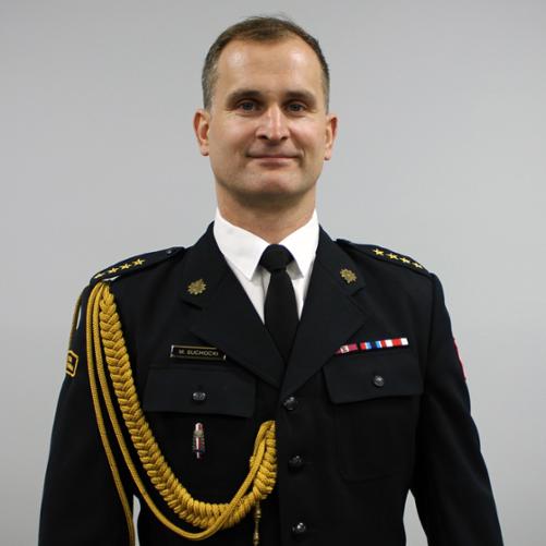 Na zdjęciu widać mężczyznę w eleganckim, galowym mundurze strażackim. Stoi na wprost obiektywu, w wyprostowanej, oficjalnej pozie. Mundur jest ciemny, z pagonami na ramionach, ozdobnym złotym sznurem na prawej stronie oraz kilkoma odznaczeniami i naszywkami. Na piersi znajduje się plakietka z nazwiskiem oraz niewielkie przypinki i barety świadczące o ukończonych szkoleniach lub wyróżnieniach. Mężczyzna ma krótkie, schludnie uczesane włosy i delikatny uśmiech. Tło jest jednolite, jasnoszare, dzięki czemu cała uwaga skupia się na postaci w mundurze