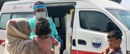 przed ambulansem lekarz mierzy temperaturę kobiecie z dzieckiem na ręku
