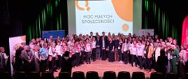 Szeroki kadr, zdjęcie grupowe. Uczestnicy konferencji pozują do wspólnego zdjęcia, stojąc razem na scenie. Za nimi widoczny ekran, na którym wyświetlona jest grafika z tytułem: Moc Małych Społeczności. Za uczestnikami, po obu stronach ekranu widoczne są także fragmenty banerów promocyjnych.