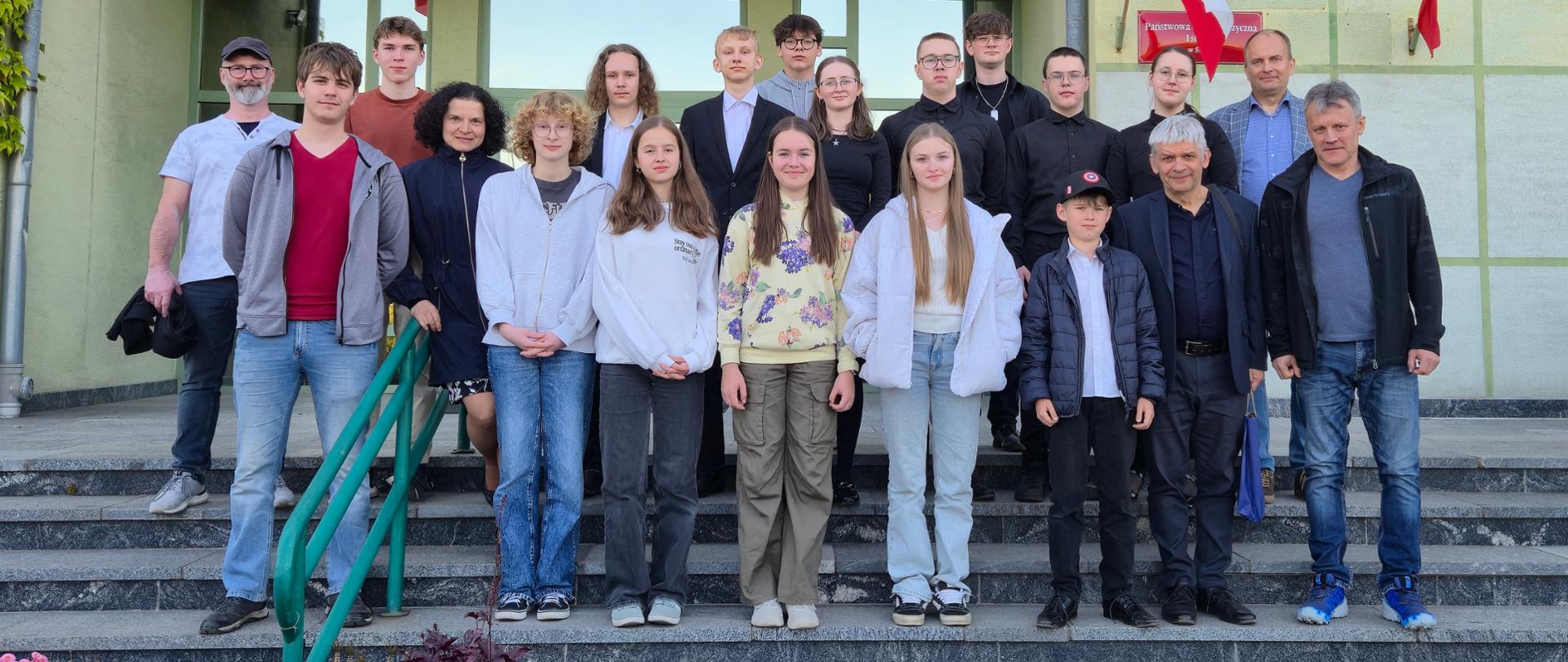grupa kilkunastu osób stojących w kilku rzędach na schodach przed budynkiem szkoły