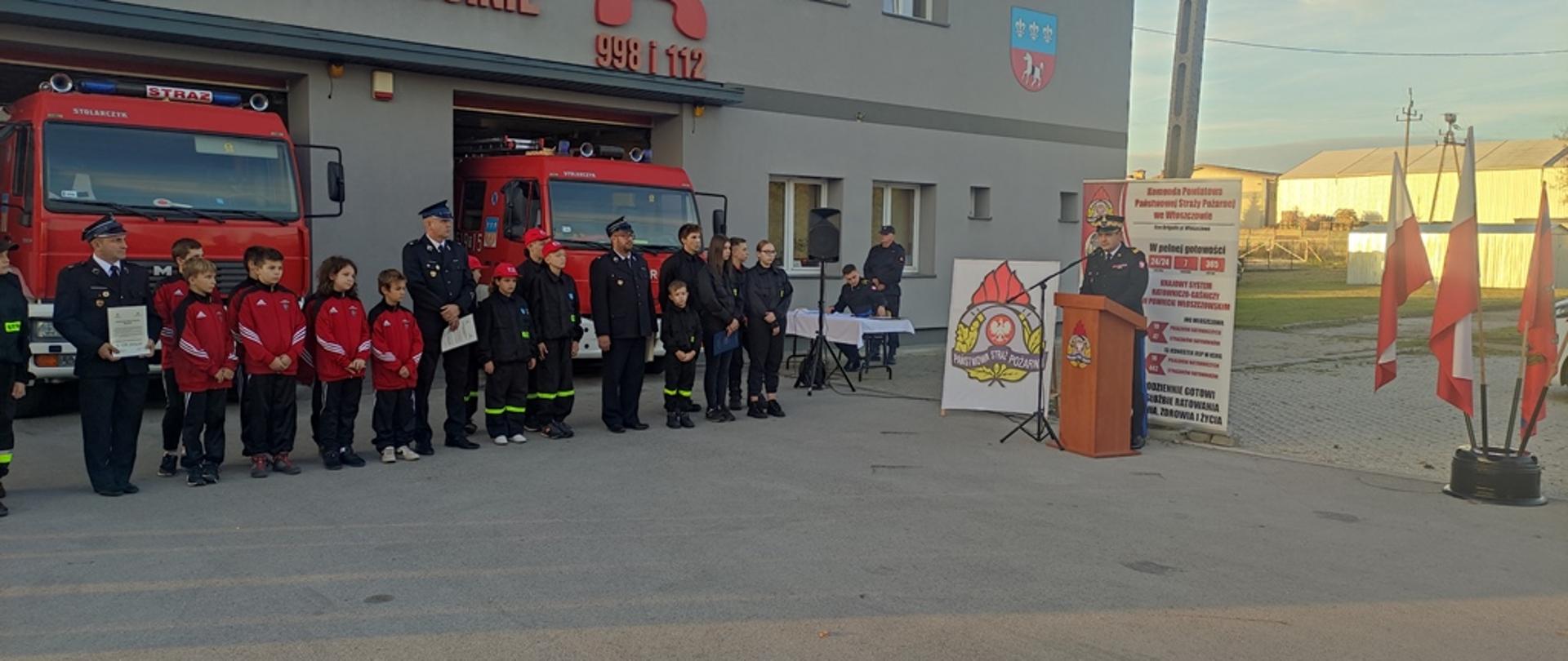 Plac przed remizą, w bramach wjazdowych do garażu ustawione samochody pożarnicze, przed samochodami młodzi strażacy wraz z opiekunami, przy mównicy komendant wojewódzki w trakcie przemówienia