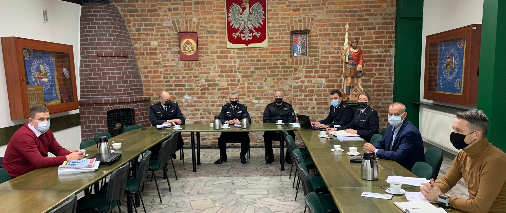 W świetlicy Komendy Wojewódzkiej Państwowej Straży Pożarnej w Toruniu przy stole ustawionym w podkowę siedzą uczestnicy spotkania: przedstawiciele Komendy Wojewódzkiej PSP w Toruniu oraz przedstawiciele związków zawodowych. Ze względu na panującą pandemie koronawirusa pomiędzy uczestnikami są spore odstępy, a każdy z nich ma na twarzy maseczkę. Przedstawiciele związków to trzej mężczyźni w cywilnych ubraniach, pięciu przedstawicieli Komendy Wojewódzkiej PSP w Toruniu, to czterej mężczyźni i jedna kobieta w mundurach.