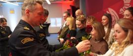 zastępca komendanta wojewódzkiego psp wręcza różę kobiecie- blondynka w białym sweterku, w tle inne kobiety