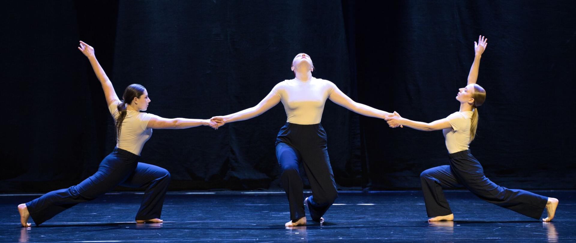 Na zdjęciu są trzy słuchaczka Studium, która występuje na scenie, na tle czarnych kotar. Są w tanecznej pozycji i trzymają się za ręce. 