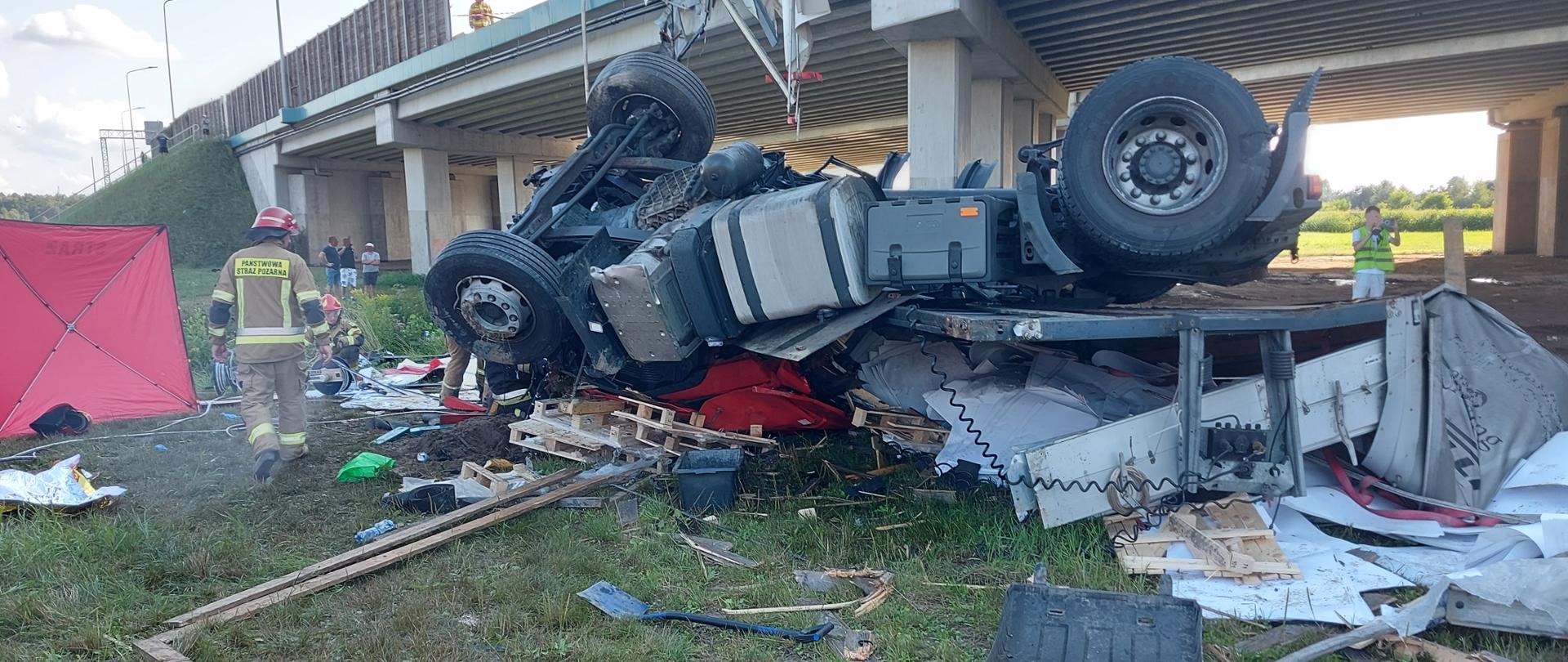 Zdjęcie pokazuje wywrócony kołami do góry ciągnik siodłowy ciężarówki, która spadła z wiaduktu z trasy S8. Na pierwszym planie widać leżące na trawie elementy samochodu ciężarowego i ładunku, które uległy zniszczeniu po upadku ciężarówki z wiaduktu: karoseria kabiny, błotnik od koła, palety, deski, a także sprzęt przysposobiony do działań ratowniczych: szpadel i kuweta na cieknące paliwo z baku. Po prawej stronie zdjęcia widać część czerwonego parawanu strażackiego. Obok parawanu jest strażak ustawiony tyłem w ubraniu specjalnym piaskowym i czerwonym hełmie. W tle za strażakiem widać klęczącego drugiego strażaka operującego przewodami hydraulicznymi na bębnie oraz osoby postronne stojące za rzeką płynącą pod wiaduktem. W centrum zdjęcia usytuowany jest zniszczony, leżący na dachu ciągnik siodłowy. Jego czerwona kabina została zgnieciona pod wpływem ciężaru naczepy, której przód widać pod tylnymi kołami ciągnika siodłowego. Za leżącym ciągnikiem siodłowym usytuowany jest wiadukt trasy S8. Na górze zdjęcia widać drewniane panele odgradzające jezdnię trasy S8, a także odcinek brakujący, który zniszczyła ciężarówka. Na wiadukcie widać stojącego strażaka w ubraniu specjalnym piaskowym i czerwonym hełmie. W tle zdjęcia widać konstrukcje wiaduktu od dołu oraz nasyp pod wiaduktem obrośnięty trawą. Po prawej stronie zdjęcia obok tylnych kół, leżącego ciągnika siodłowego, stoi osoba w kamizelce odblaskowej, a za nią widać łąkę i las.