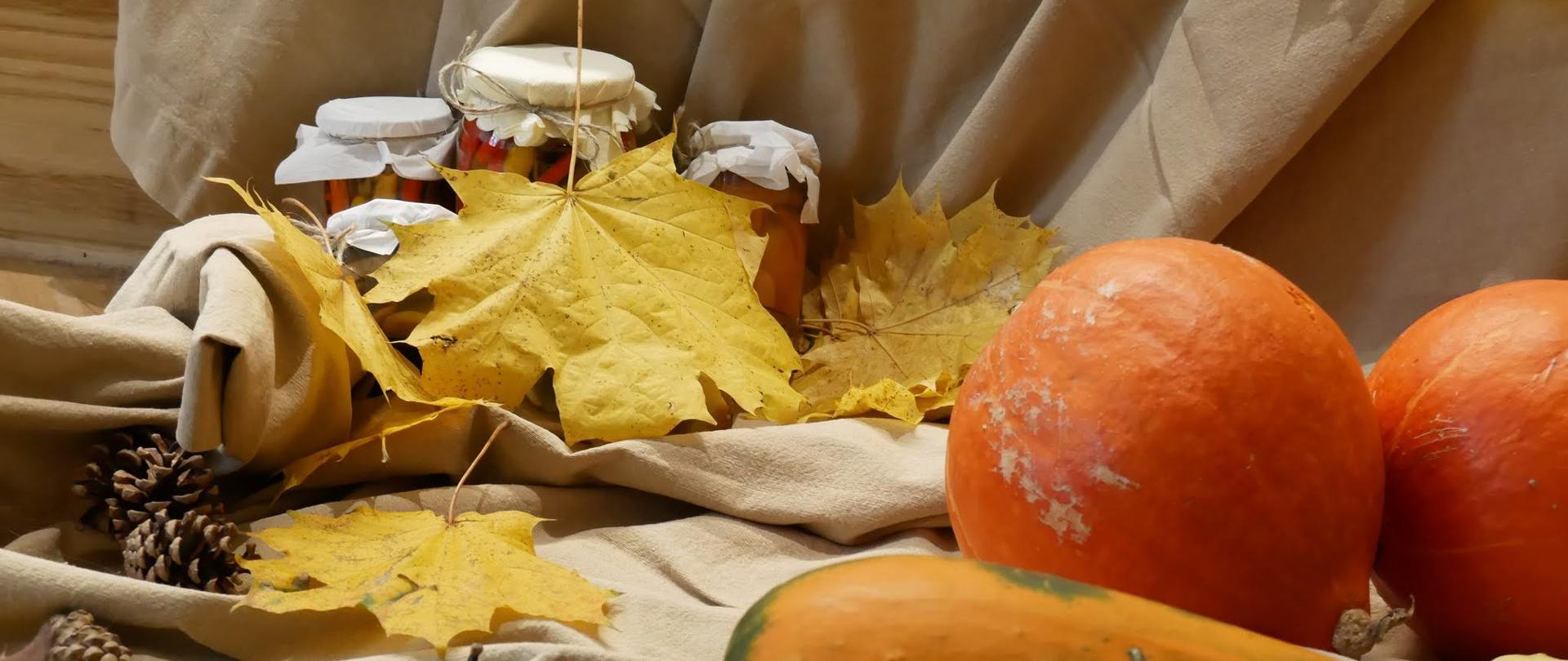 Na beżowym materiale ułożone, dynie, szyszki, jesienne liście oraz słoiczki z przetworami.