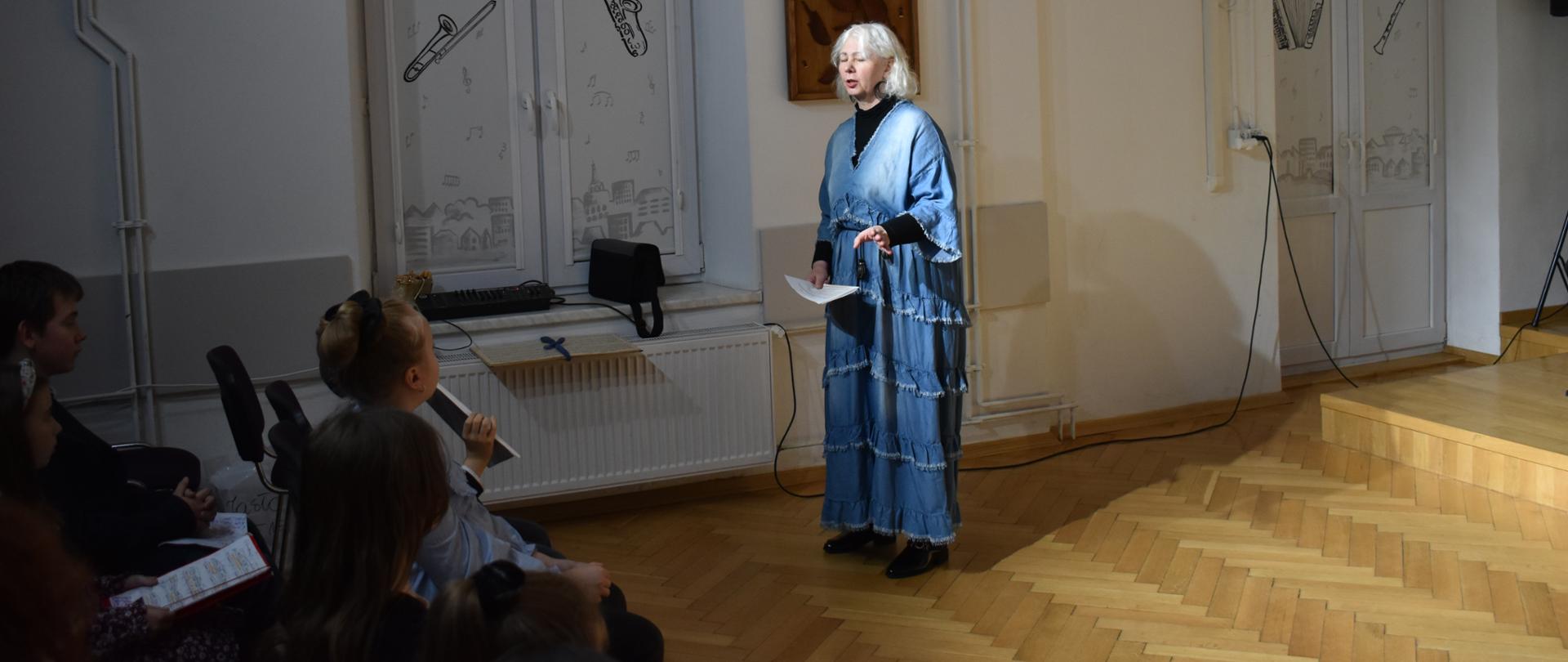 Z lewej strony na pierwszym planie widać głowy siedzących dzieci patrzących w stronę kobiety ubranej w niebieską sukienkę trzymającą w ręku kartkę. Za kobietą na ścianie wisi praca przedstawiająca rozbite skrzypce.
W tle biała ściana i zasłonięte na biało okna na których znajdują się kontury instrumentów i pięciolicie pięciolinie z nutami.
