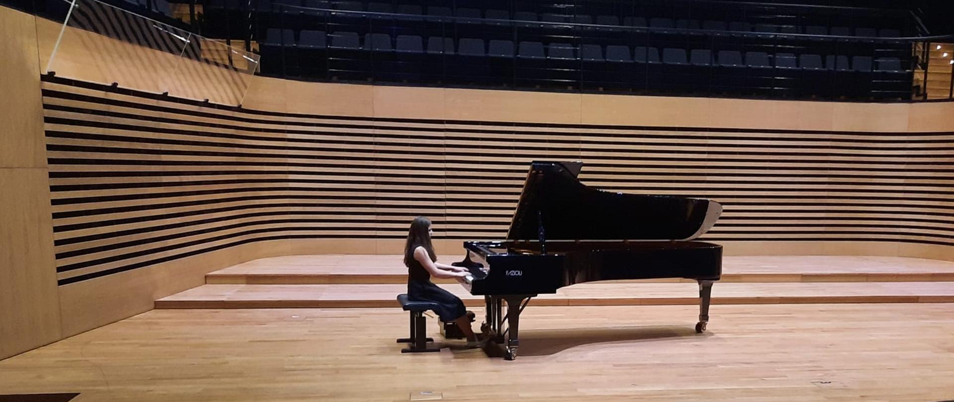 Nastolatka gra na fortepianie w sali koncertowej, z przodu widać od tyłu fragment widowni z publicznością. Po prawej stronie estrady stoją organy.