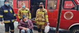 Troje strażaków w mundurach prowadzi do punktu szczepień starszego mężczyznę na wózku inwalidzkim. Wszystkie osoby są posiadają maseczki na twarzy. W tle widać czerwony bus strażacki z OSP w Samborcu. 