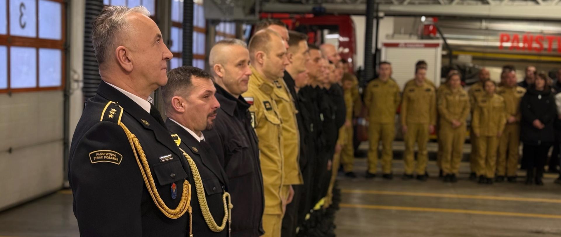 Zdjęcie wykonane z boku szeregu strażaków stojących w postawie zasadniczej. Na pierwszym planie widać profile dwóch oficerów w ciemnogranatowych mundurach wyjściowych ze złotymi sznurami galowymi. Za nimi stoją kolejni strażacy w piaskowych mundurach. Perspektywa prowadzi w głąb garażu, gdzie widać białe bramy wjazdowe z numerami.