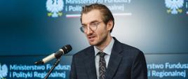 Wiceminister funduszy i polityki regionalnej Jan Szyszko na konferencji prasowej w MFiPR nt. odblokowania środków dla Polski z Funduszy Europejskich, wiceminister stoi przy pulpicie i przemawia do mikrofonu, za jego plecami napis MFiPR