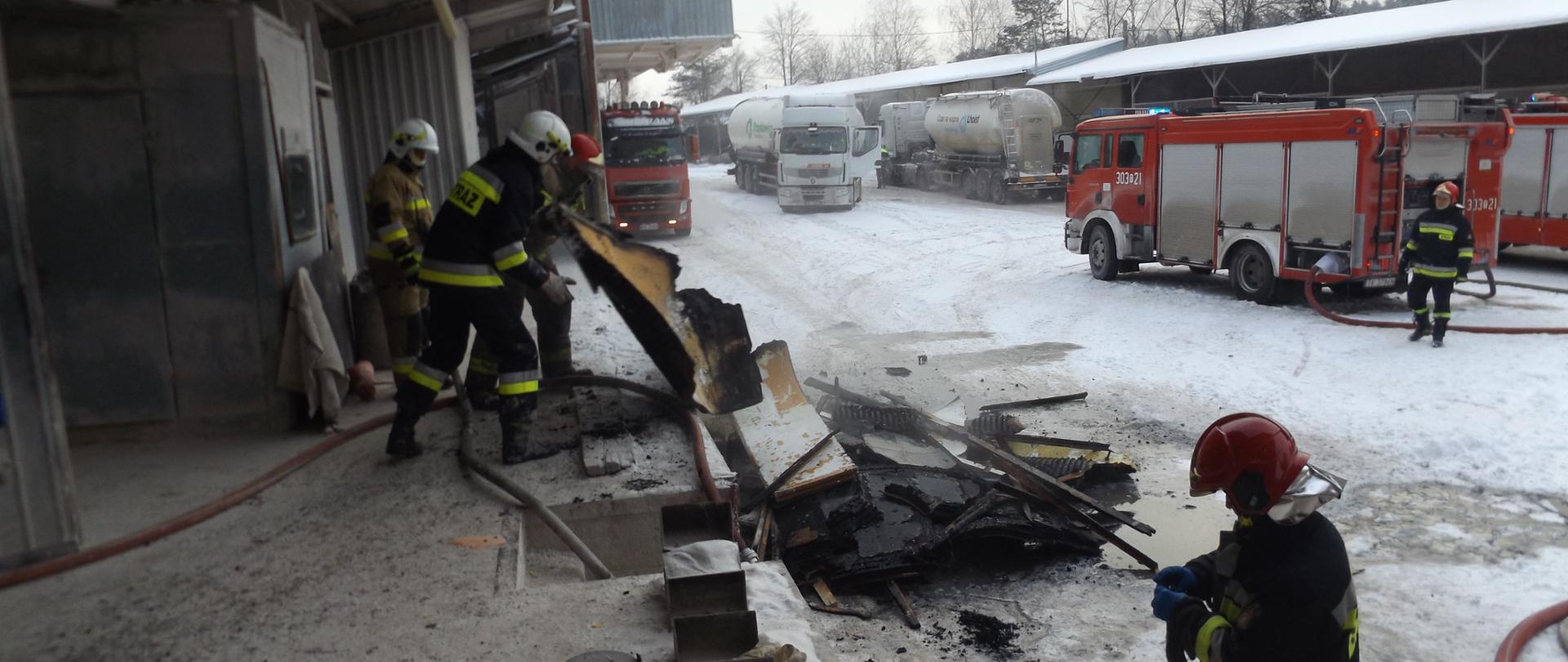 Na zdjęciu znajdują się strażacy porządkujący teren akcji. Przed budynkiem nadpalone wyposażenie warsztatu. W tle znajdują się samochody strażaków oraz pozostała część zakładu.