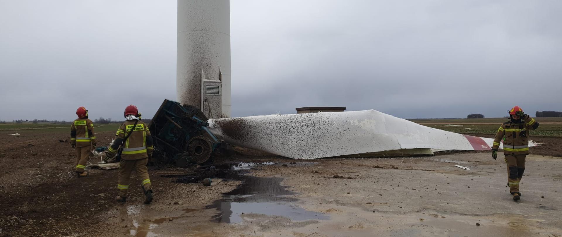 na zdjęciu trzech strażaków w czasie interwencji na polu stojący przy uszkodzonej elektrowni wiatrowej na ziemi leży jedna z łopat wiatraka